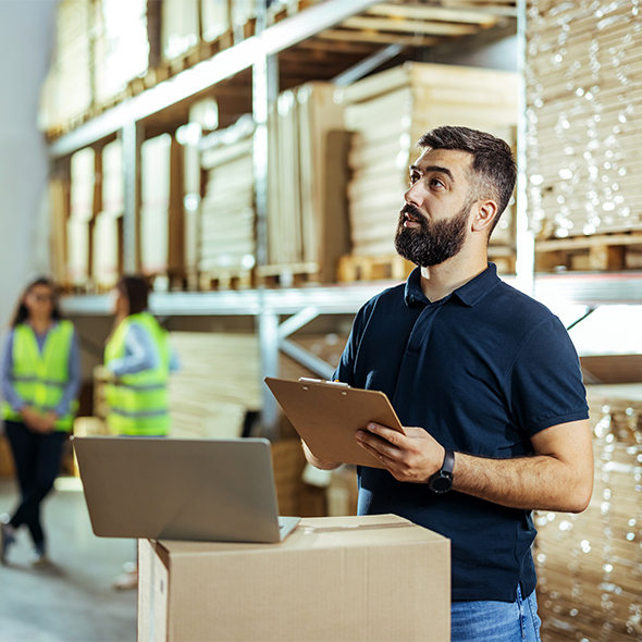 worker in warehouse looking up with a clipboard and laptop 