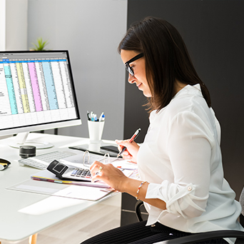 woman working at a desk operating a calculator while writing on a notepad in front of a computer